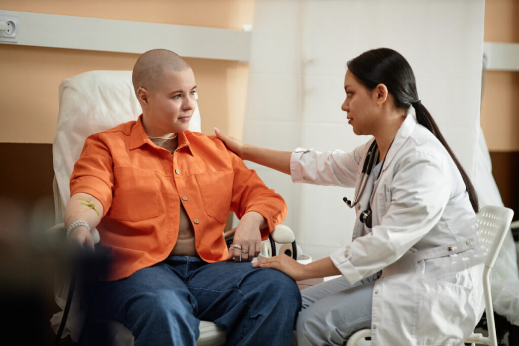 Side view portrait of young nurse comforting cancer patient receiving chemotherapy treatment in hospital. More Young Adults Are Being Diagnosed with Cancer 